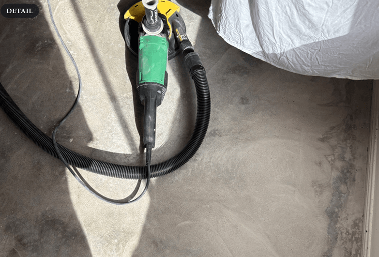 The concrete slab being mechanically ground with a vacuum-shrouded grinder to strip staining and the weak toplayer so the waterproofing bonds to sound concrete