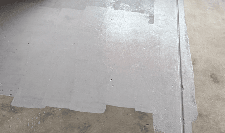 The first coat of epoxy waterproofing membrane going down over the prepared slab, with bare concrete still visible alongside and the joint on the right also coated.